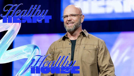 A smiling bald man with glasses and a beard stands on a stage with a blue background. Blue text overlay reads, 'Healthy Heart, Healthy House.'